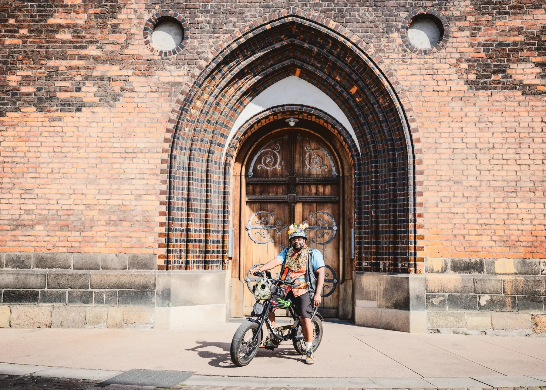 En stor gotisk murstensbue og en trædør til en kirke fylder billedet; FatbikeHero står lille foran med sin cykel.