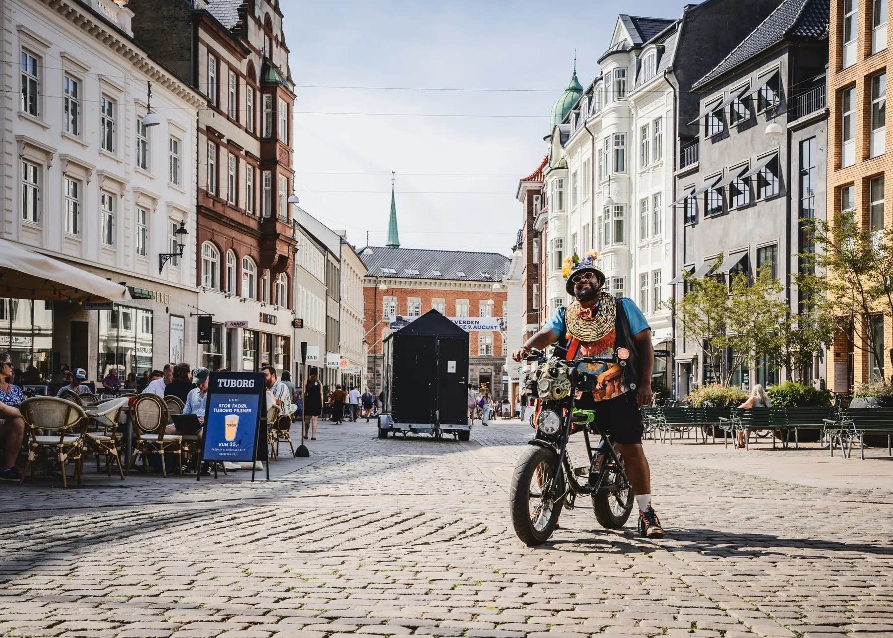 Vidt udsnit af en brolagt plads i Aarhus med cafeborde og et ølskilt; FatbikeHero sidder på sin cykel i højre side af billedet.