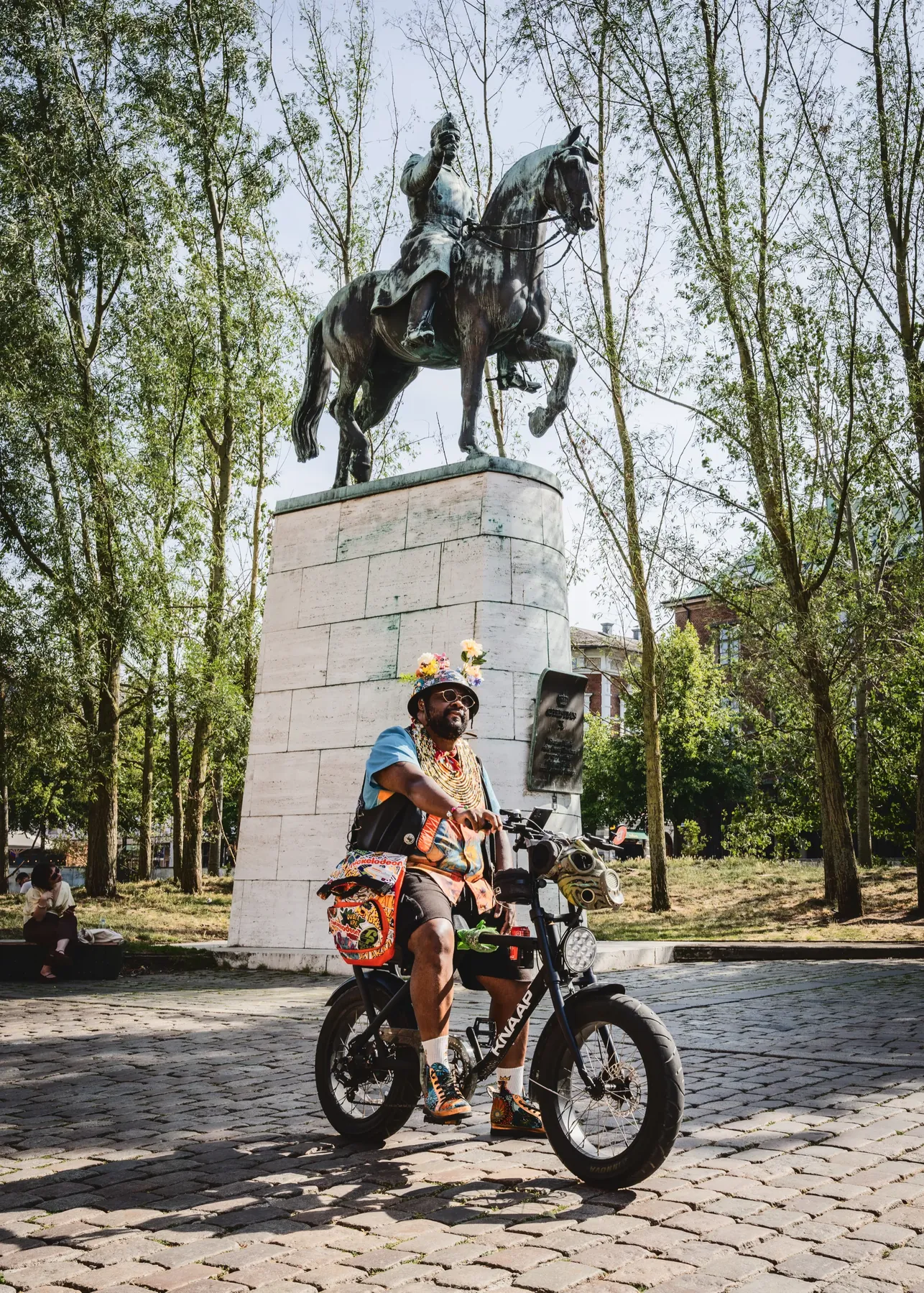 FatbikeHero på sin fatbike under en bronzerytterstatue på en høj stensokkel omgivet af træer.