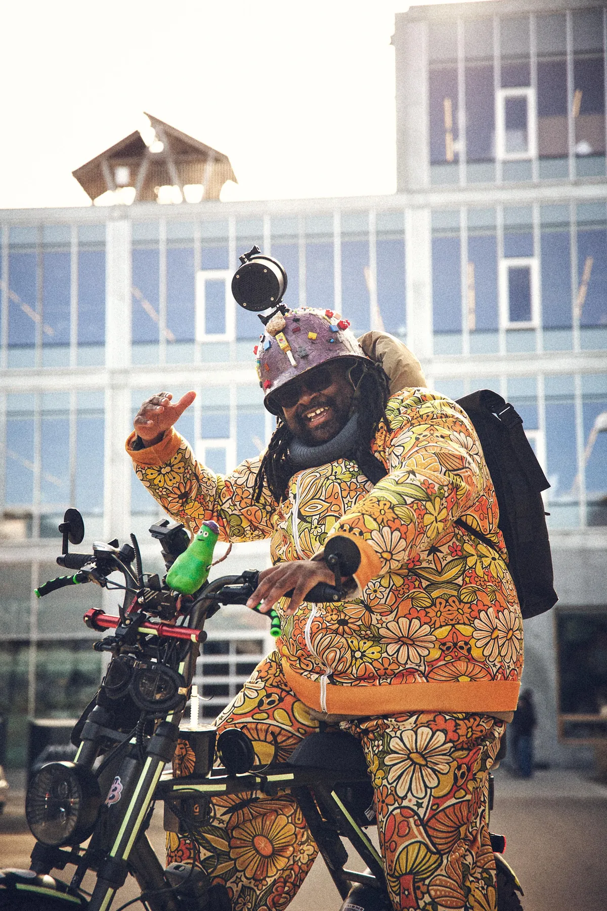 FatbikeHero på cyklen i en helt blomstret dragt med den ene hånd i vejret og modlys bag sig.
