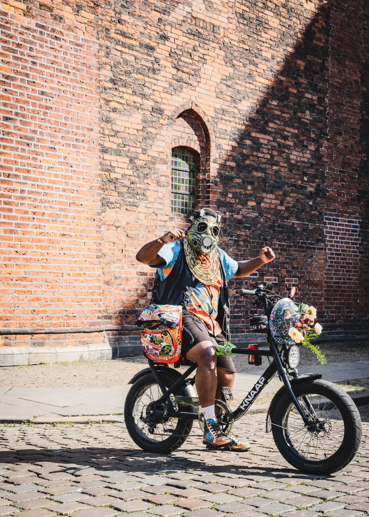FatbikeHero i malet gasmaske og bred stofkrave med begge hænder i vejret på en sort KNAAP-fatbike foran en kirkemur i Aarhus.