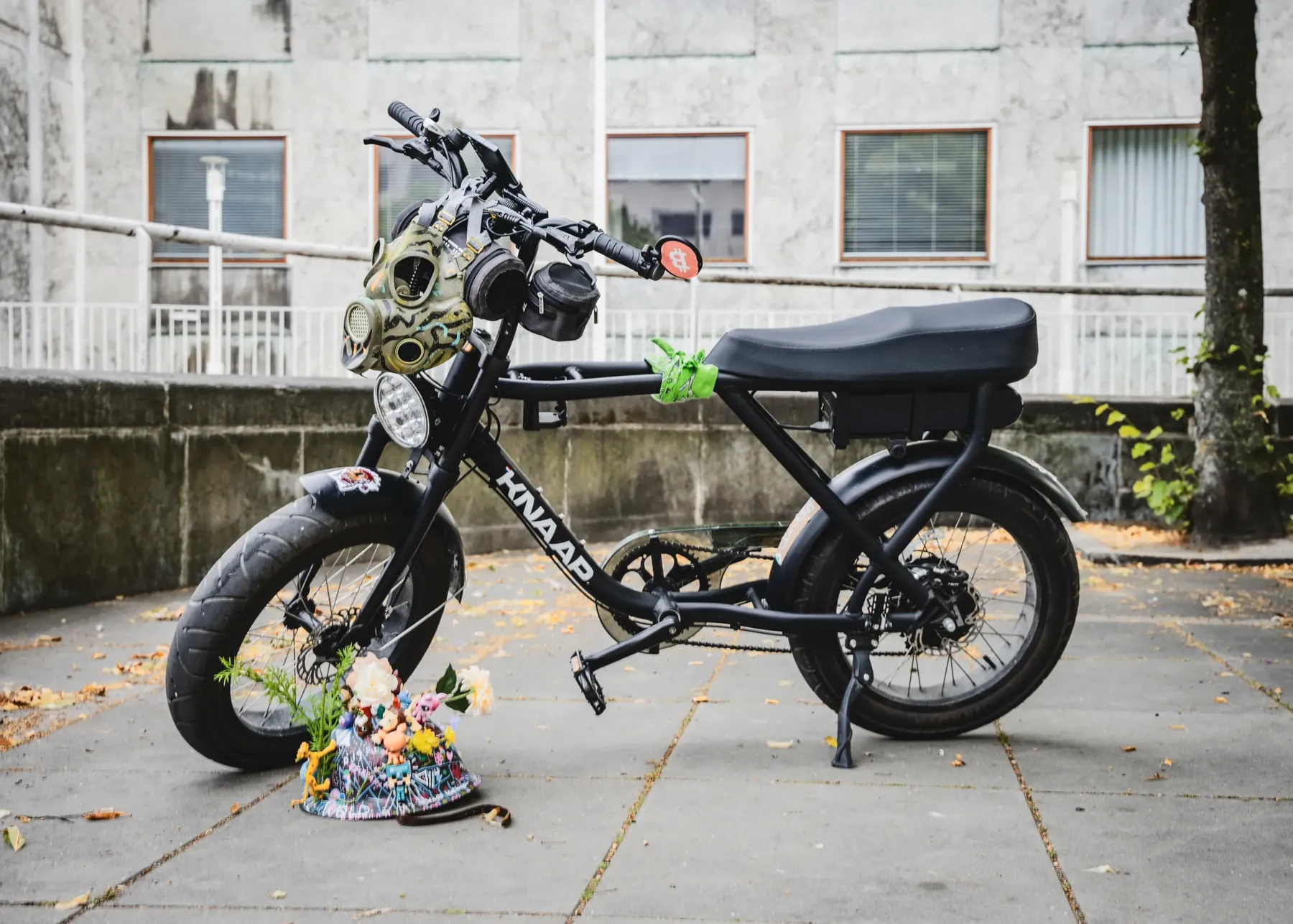 Den sorte KNAAP-fatbike parkeret alene på et fortov med gasmasken hængende på styret og blomsterhjelmen stillet på jorden.