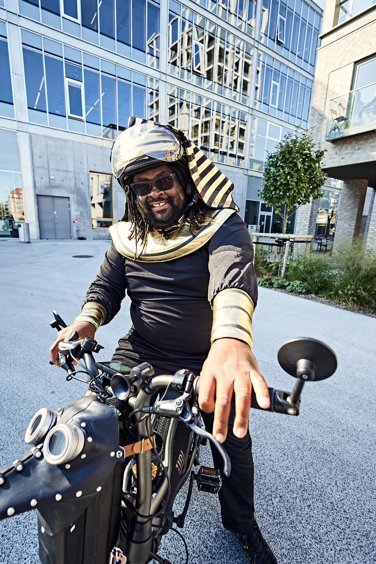 Vidvinkelbillede taget forfra fra cyklen; FatbikeHero i faraokostume rækker den ene hånd mod linsen.
