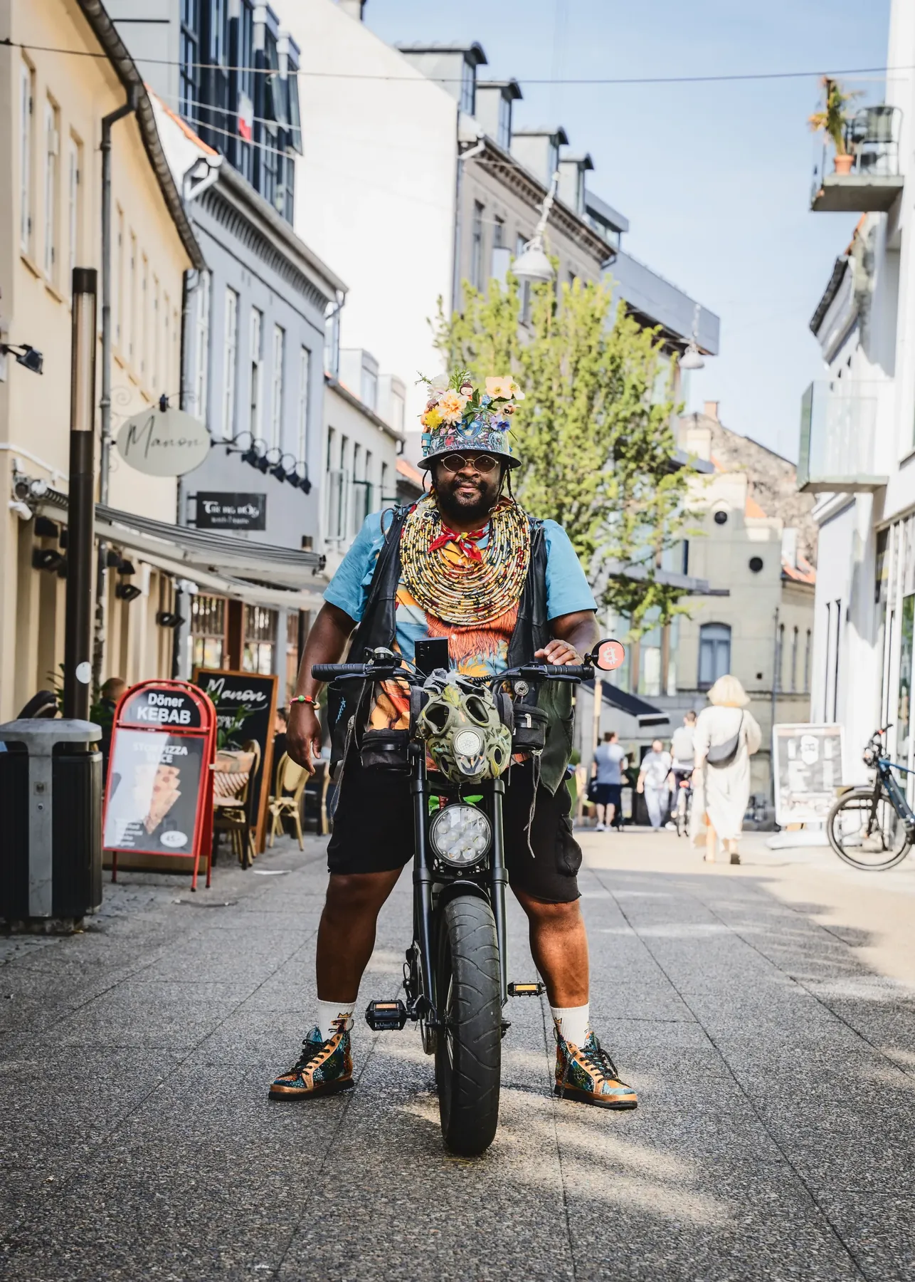Helfigursportræt af FatbikeHero stående over sin fatbike i en smal handelsgade i Aarhus med caféskilte bag sig.