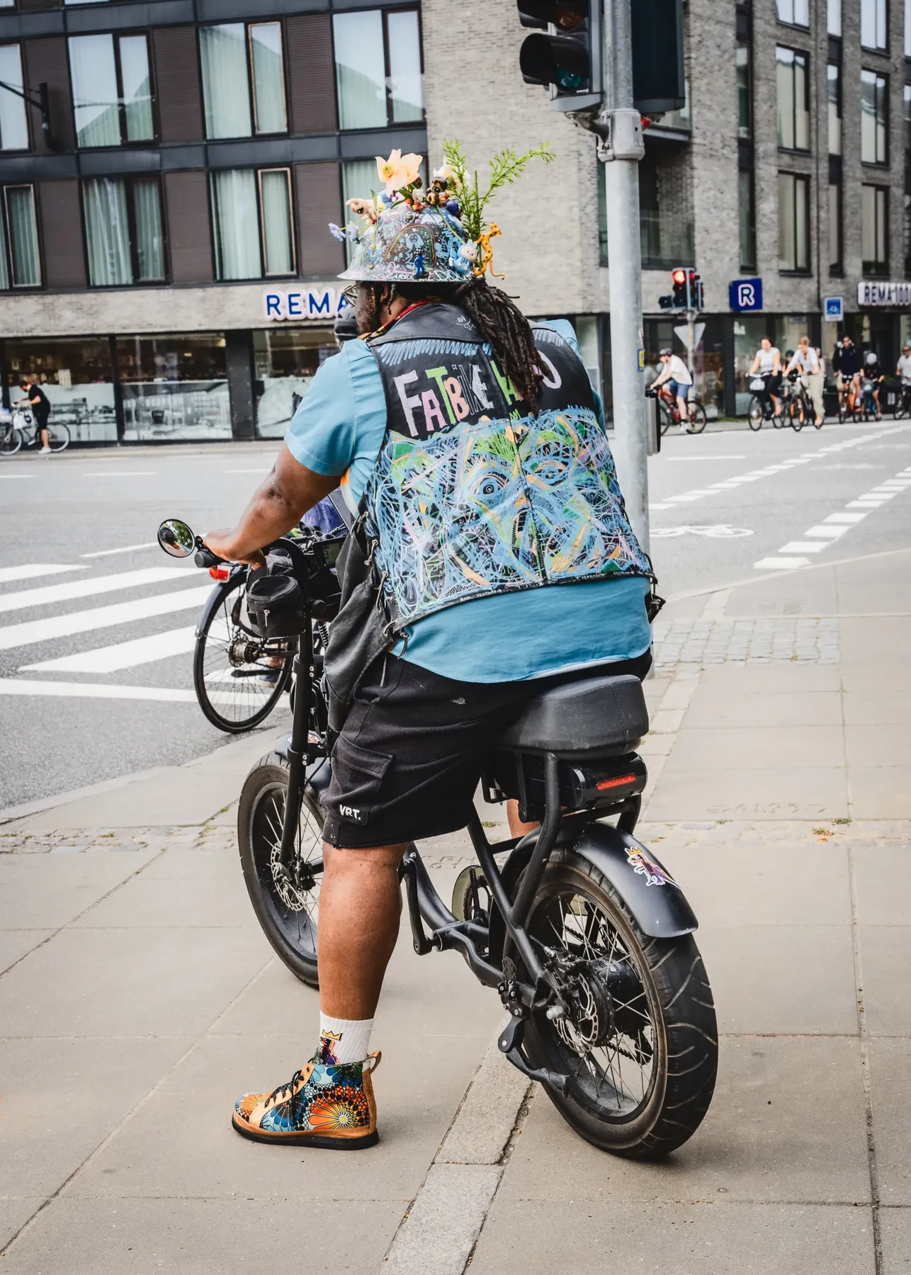 Set bagfra ved et kryds: den håndmalede FATBIKEHERO-vest, dreadlocks og blomsterhjelmen, mens der ventes på rødt.